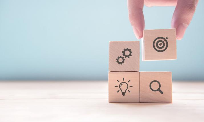Hand arranging wooden blocks with the following icons: two cogwheels, a dart board, a lightbulb and a magnifying glass